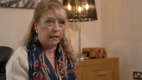 Denise Fergus, who has shoulder length blonde hair, and is wearing a white blouse and a scarf with a floral pattern, sits on a sofa and speaks to the camera