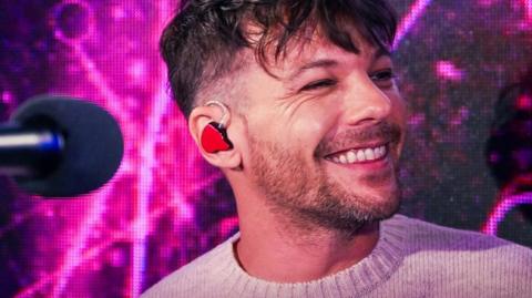 Louis Tomlinson smiles behind a mic. He has short brown hair and wears a beige jumper