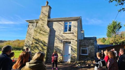 Councillor Taylor gives a speech to a crowd of people outside a grey brick farmhouse which has blue ribbon stretched across the front door.