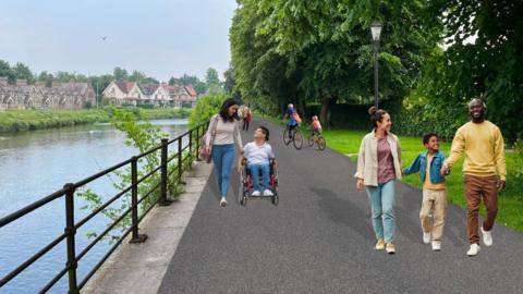A CGI image of what the riverside path could look like. The tarmacked path has pedestrians, cyclists and a wheelchair user of different ages using the path. There is a black metal banister on the edge of the water on one side of the path, while the other has a grass strip with trees. Over the water there are houses.