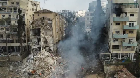 rubble from an iranian strike is show. smoke is rising over it and men in orange high vis can be seen working through the damaged site. it is a zoomed out photo showing what appears to be a residential street.