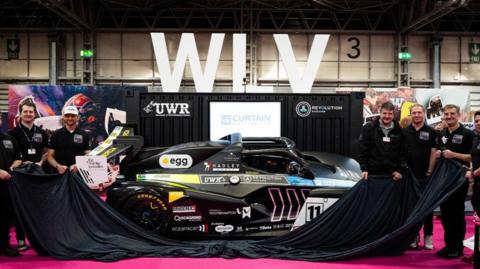 A group of men smiling and wearing black t-shirts are holding a black cloth unveiling a black racing car.