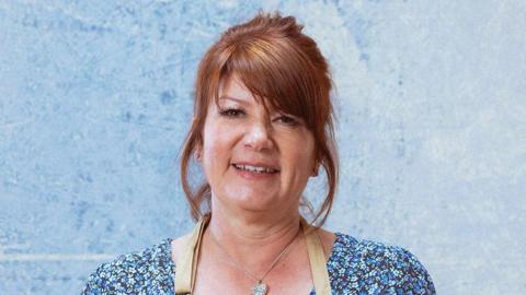 A woman with ginger hair smiling at the camera. She is wearing a blue floral top and is stood in front of a like blue background
