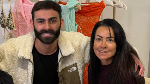 A man in a beige fleece and black t-shirt with his arm around a woman with black hair. Both are sat behind a clothes rail.