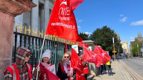A number of people are standing in front of railings at the entrance to a museum. They are dressed warmly and waving red banners with the logo of Unite, the union, on them.