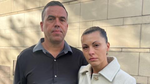 John and Jane Brady stand outside court. They are looking straight into the camera. John is taller than his wife. He has brown hair and is wearing a shirt. Jane has her brown hair pulled back. She's wearing a white coat with gold buttons.