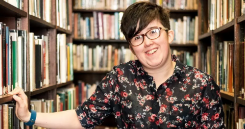 Lyra McKee is wearing a black and red floral shirt.
She is standing in front of bookcases in a library.
Her hand is rested on a shelf and she is smiling.