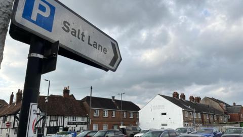 A pointed road sign directed at a car park under a cloudy sky. The sign says "Salt Lane". The tops of cars can be seen and terraced buildings are in the background.