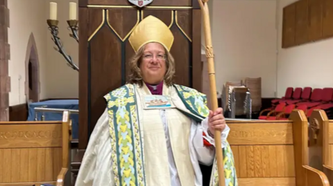 Tricia Hillas, she wears a decorated white robe and golf mitre and holds a gold staff while standing in front of a grand wooden chair.