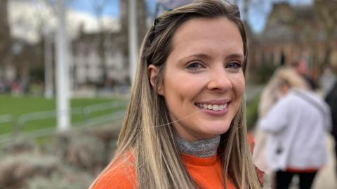 A woman wearing an orange top smiles into the camera. She has blonde hair past her shoulders and stands next to a stone wall with grass behind it.