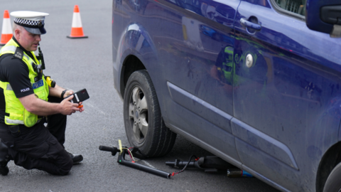 A police man kneels next to a blue van, which has a broken e-scooter underneath it. 