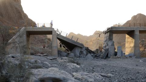 A damaged bridge with people standing on the remaining structure with mountains in the background, and rubble and rocks in the foreground.