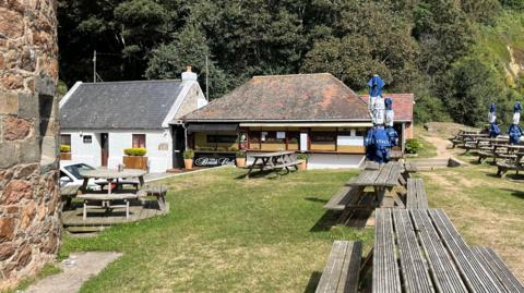 The Fermain Beach Café. A small white and brick orange building with a Martrello Tower on the left, with a number of picnic benches in front of it. 