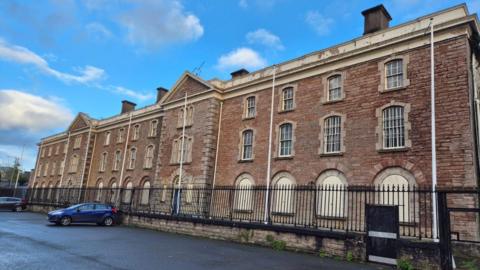 The front view of Armagh Gaol on a sunny spring evening. The three-story, 14-bay Georgian building is surrounded by a low perimeter wall, topped by a decorative, spiked iron fence. The windows and doorways are arched. The ground floor windows are boarded up, while upper floor windows are secured by white metal bars. There are two hatchback cars parked on the pavement in front of the gaol. 