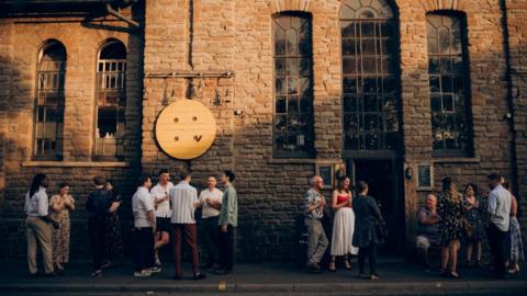 A crowd of people stand outside a converted church building which has tall windows and light coloured stonework.