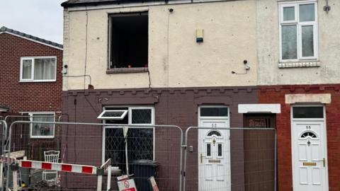 An end terrace house with top window blown out of the frame, and metal and safety barriers surrounding the ground floor exterior on the pavement