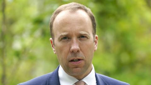A man standing outside. He has thin light brown hair and is frowning slightly with his mouth open. He is wearing a blue suit with a white shirt and a light brown tie.