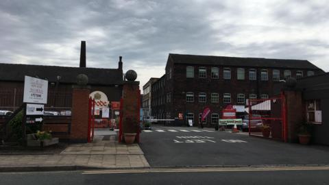 The outside of two brick buildings. One on the right has lots of small windows and sits behind an open red gate. On the left, the building has a large, long chimney and a white sign depicting a gold crown and the word "Spode". There is a white sign in front of it that reads "Spode works, a potbank"