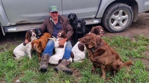 Craig sits beside a silver vehicle and cuddles eight dogs.