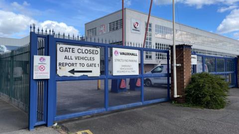 The closed blue gates outside the Vauxhall plant. A grey building with many windows and a Vauxhall logo is behind the gates.