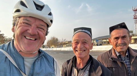 Stephen with two men smiling wearing traditional caps by the side of a road. Stephen is wearing a white cycling helmet and a lightweight blue jacket. 