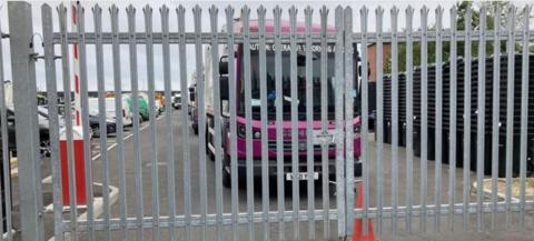 A purple waste lorry sits behind metal gates. An orange cone can be seen in front.