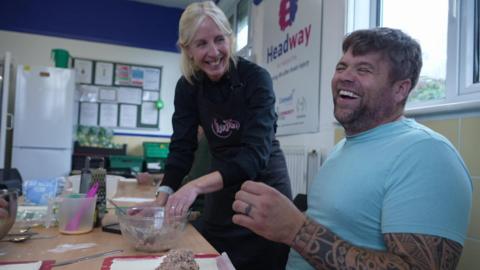 A man wearing a blue T-shirt laughing and smiling with a lady with blonde hair. The man has tattoos up his arm. The woman is wearing a black apron.