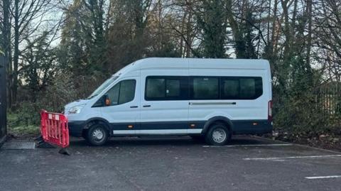 A white minibus is parked in a car park with a red plastic fence placed in front of it. The car park is surrounded by a mental fence with vegetation woven around it.