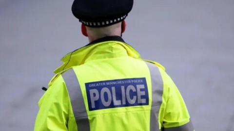 A male police officer from Greater Manchester Police on duty in the city centre. They are facing away, wearing a yellow high-vis jacket and a black police hat.