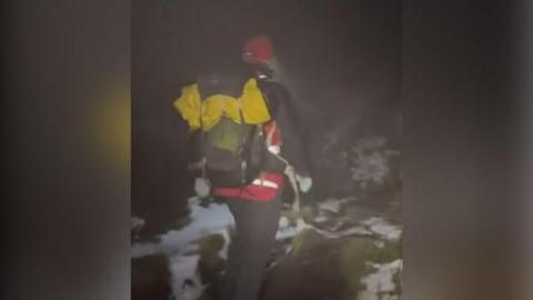 Mountain rescue team volunteer in climbing gear walking in wintry conditions with snow on the hills. He is pictured from behind. It is dark and raining. He has a red hi vis coat and a yellow equipment at the top of his green and grey ruck sack. 