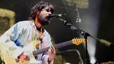 A bearded man is holding an acoustic guitar and singing into a microphone on a stand at Reading Festival.