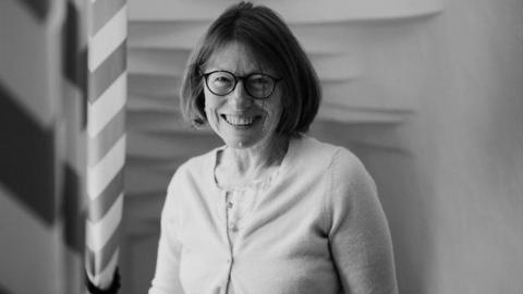 A black and white photo shows Jane Henshaw smiling at the camera. She is wearing dark framed glasses and has bobbed hair, with a pale coloured top and cardigan.