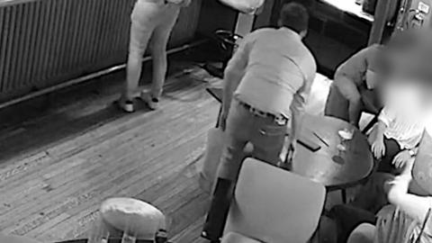 A man stood up by a table in a bar with his back to the camera.