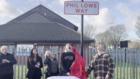 Five people stand underneath a road sign. It is in red letters against a white background and says Phil Lowe Way. Underneath are members of his family, including his son, who has just pulled down a red sheet that was covering the sign.