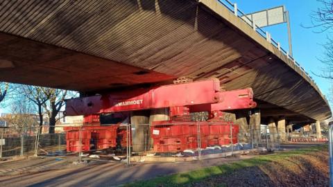 A large red metal structure props up the grey concrete flyover road. The equipment is surrounded by grey fencing.