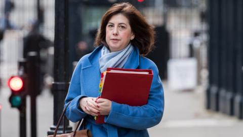 Lucy Powell seen wearing a blue jacket while holding a handbag and a red folder on the street.
