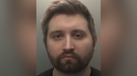 A mugshot of Declan Guess, who has short brown hair and has a beard. He is wearing a black T-shirt.