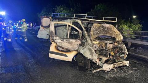 The charred remains of a white van on a slip road, next to the barrier. The front is crushed, suggesting a crash. Fire crews are gathered on the road to the left.
