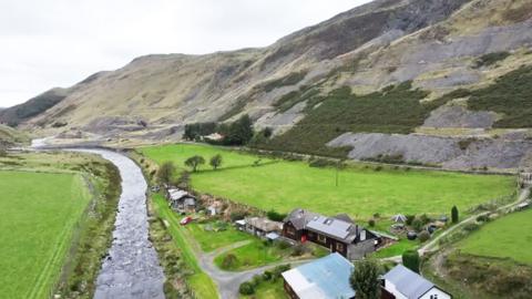 A river flowing through countryside in Wales by some hills and a few buildings.