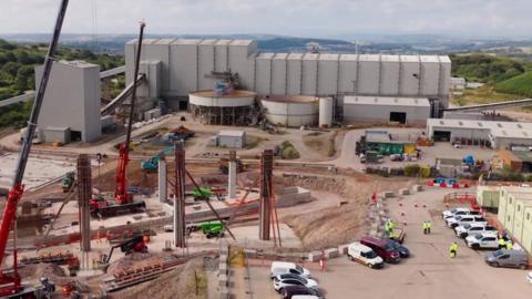 There are grey industrial buildings and huge upright structures at the mine. There are cars parked nearby and containers on the edge of the shot, with the moors in the background.