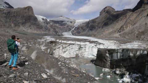 A scientist takes a photo as part of an annual monitoring project of Austrian glaciers administered by the Austrian Alpine Association in 2023. In the background the glacier slopes down the rocky mountainside, while in the foreground chunks of ice are in a stream.