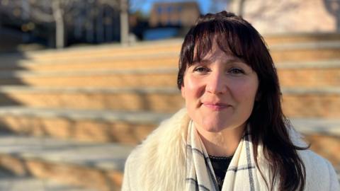 Joanna Merrett on the University of Exeter campus wearing a check scarf and a cream coat with a fluffy collar - the background is blurred.