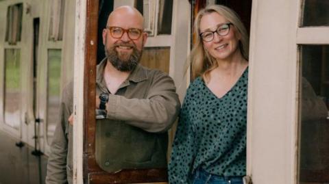 Joanne Ormesher is standing the doorway of a vintage railway carriage. She is wearing a green top with black spots and blue jeans - she has blonde hair and large dark glasses.