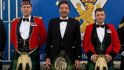 A row of men, including Jimmy Fallon, wearing kilts standing in front of a sign which reads 'His Majesty the King's Royal Guard'.