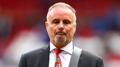 A bearded man wearing a red tie and suit inside a football stadium. He is playing a guitar