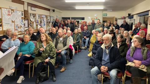 More than 50 people sitting and standing in a room facing the front to debate the future of the hospital. A woman wearing a green coat and talking while pointing her finger towards the front of the room.