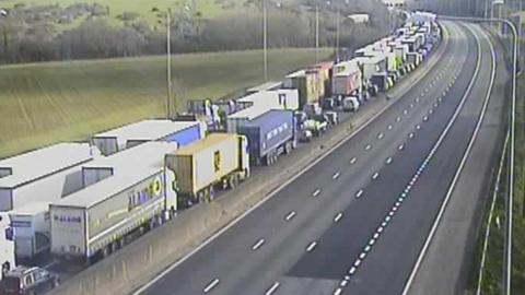 A queue of traffic made up of lorries and cars queueing on the M25. The other carriageway is completely empty.