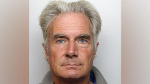 A close up of a mug shot showing an older man with grey hair and eyebrows. He has blue eyes.