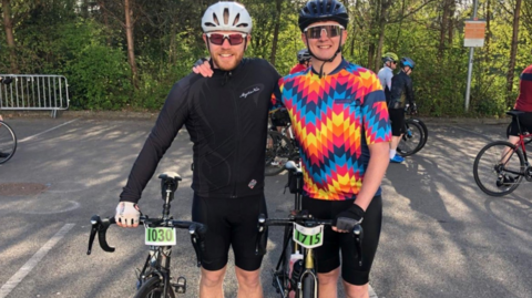 Jim and Dave are standing side by side holding their bicycles and with their arms around each other. Jim on the left is wearing black cycling gear and Dave has a multi-coloured striped t-shirt and black shorts. There are other cyclists behind them - each bike has an event number on the handlebars - and there are trees in the background
