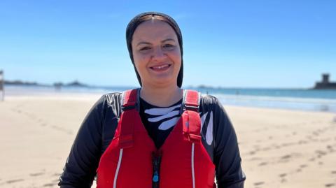 Maha is wearing a red life jacket over dark watersports clothing stands on a sandy beach facing the camera. Footprints trail across the sand toward the shoreline, with calm blue water, a surfboard in the distance, and a small offshore structure visible on the horizon beneath a clear sky.